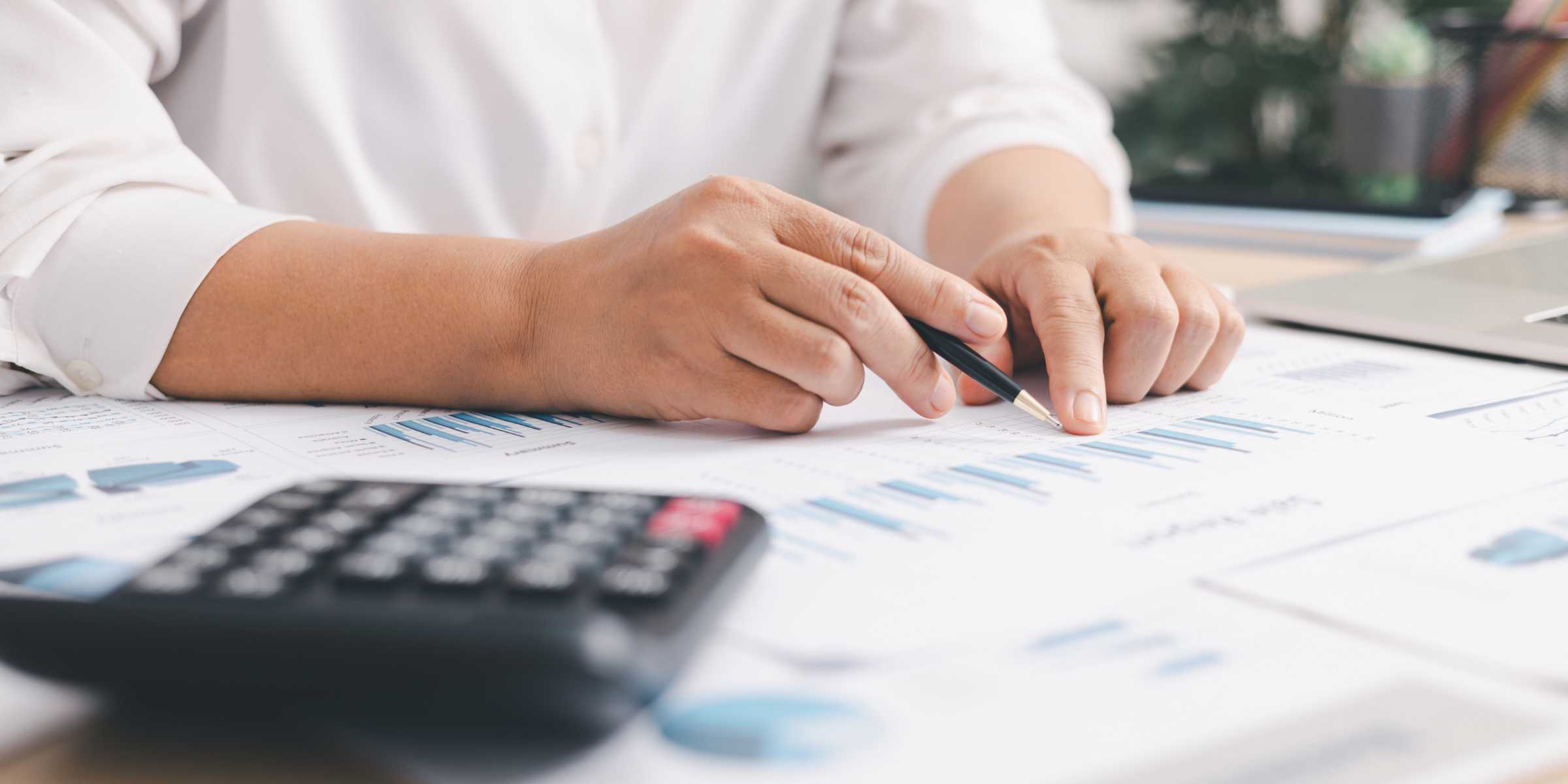 A person reviews financial charts and graphs on paper, holding a pen, with a calculator and a laptop nearby on the desk.