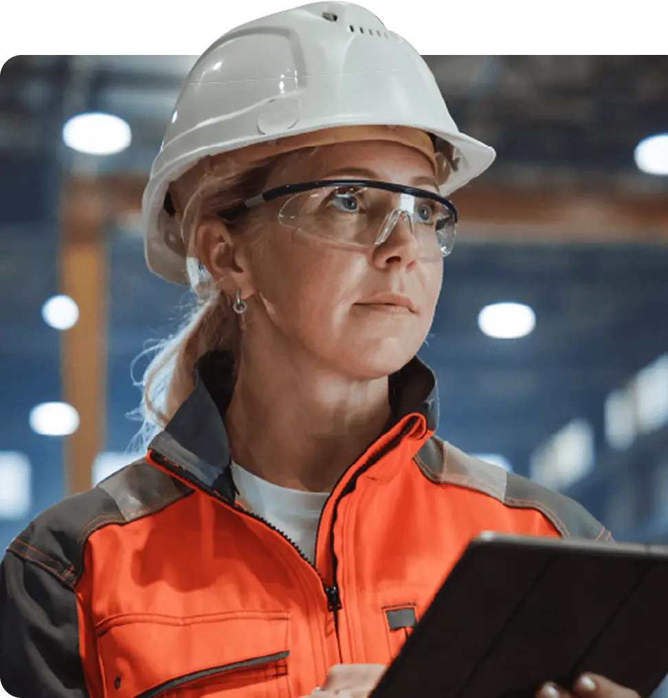 A woman in a hard hat and glasses is holding a tablet, focused on her work in a construction or engineering setting.