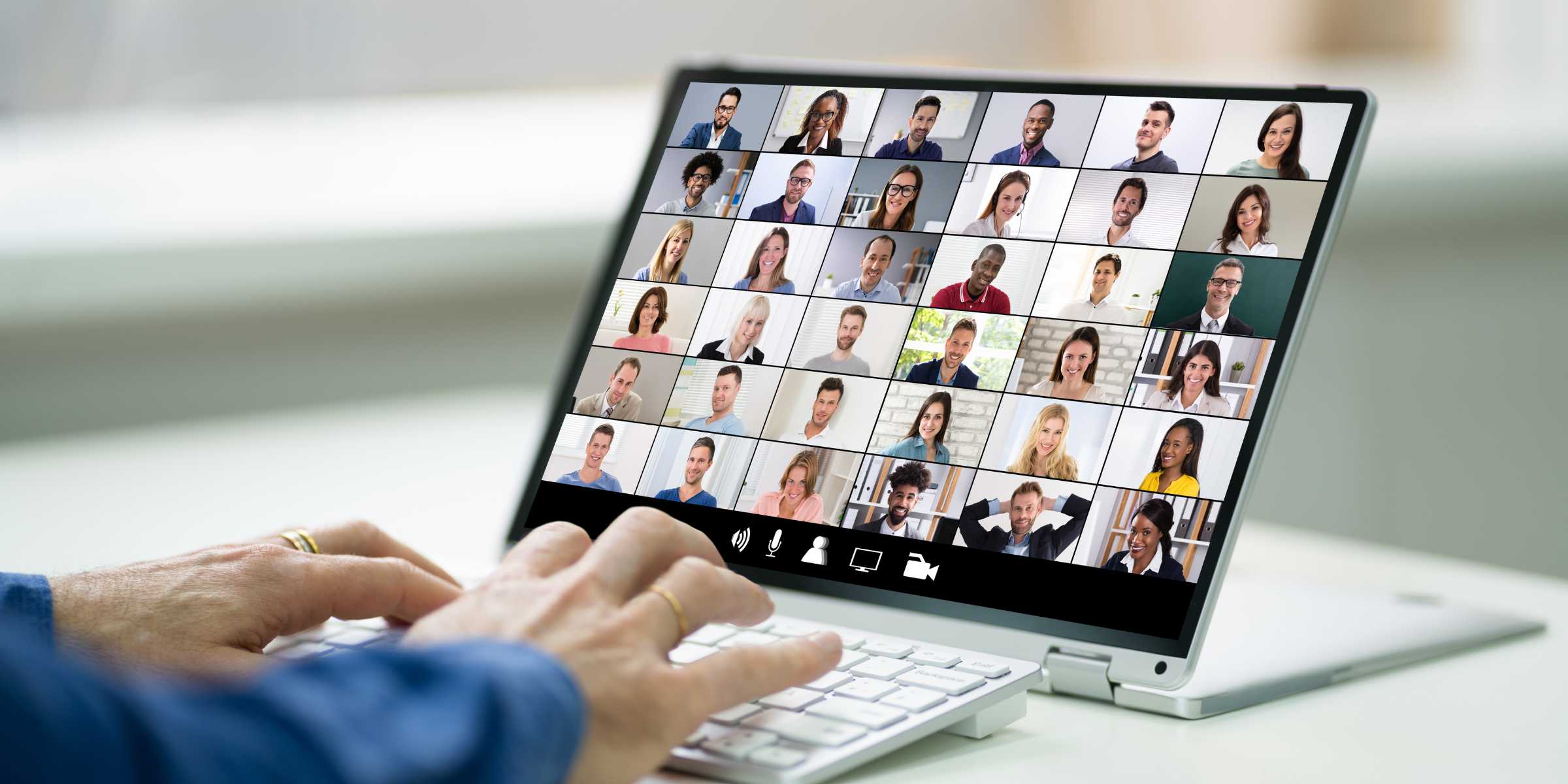 A person types on a laptop, with multiple individuals visible on the screen, engaged in a virtual meeting or discussion.