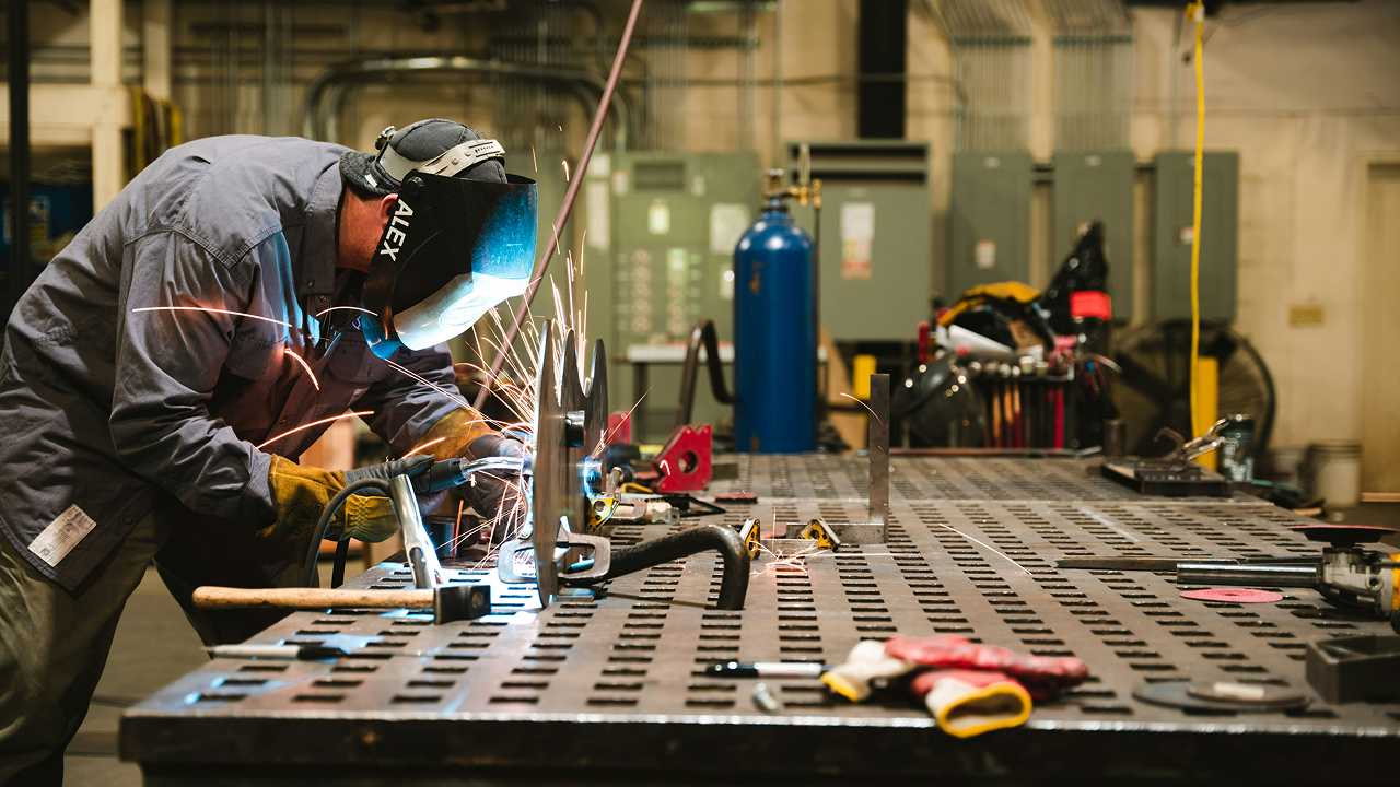 A man wearing a welding helmet is focused on welding a metal piece on a work table.
