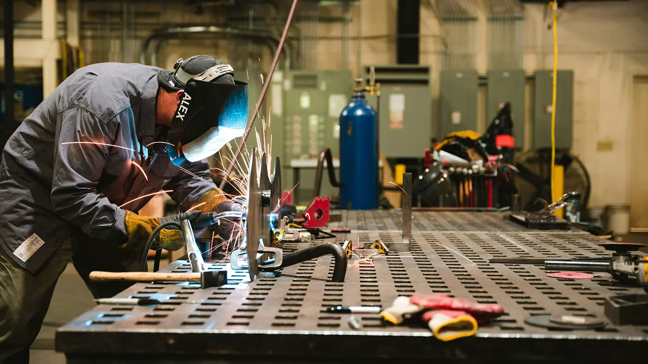 A man wearing a welding helmet is focused on welding a metal piece on a work table.