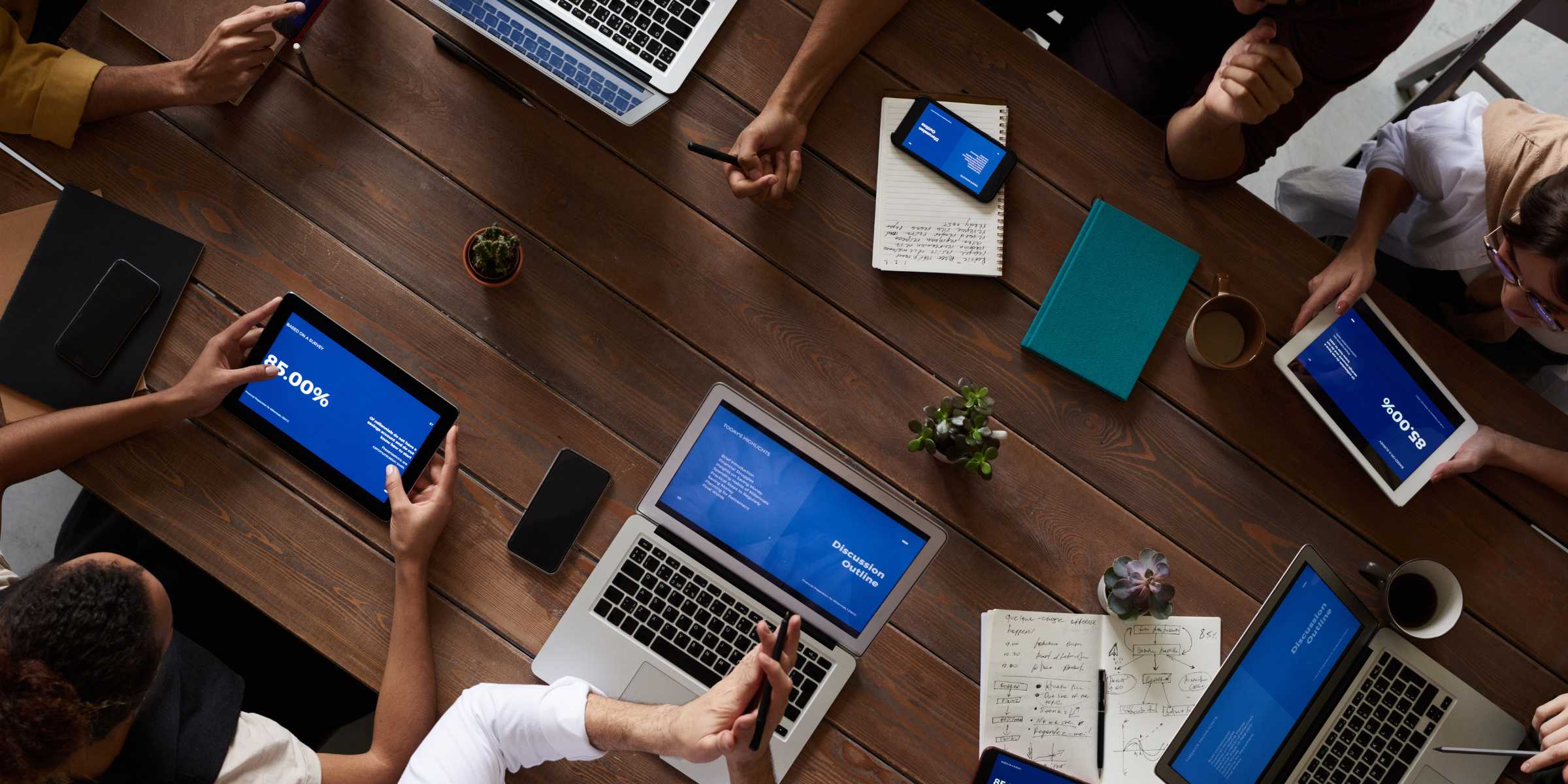 A group of people sit around a wooden table with laptops, tablets, notebooks, and coffee cups, working and discussing documents.